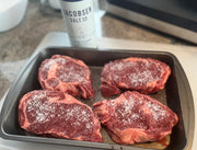 Raw Grassfed steaks seasoned with salt on a baking tray, with Jacobsen Salt & Pepper container in the background.