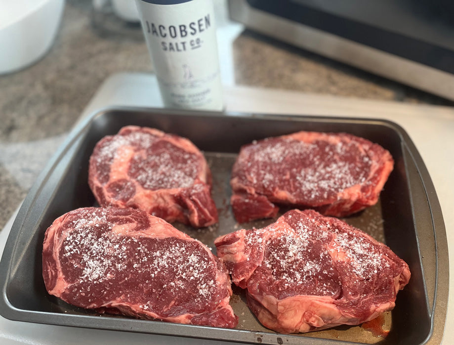 Raw Grassfed steaks seasoned with salt on a baking tray, with Jacobsen Salt & Pepper container in the background.