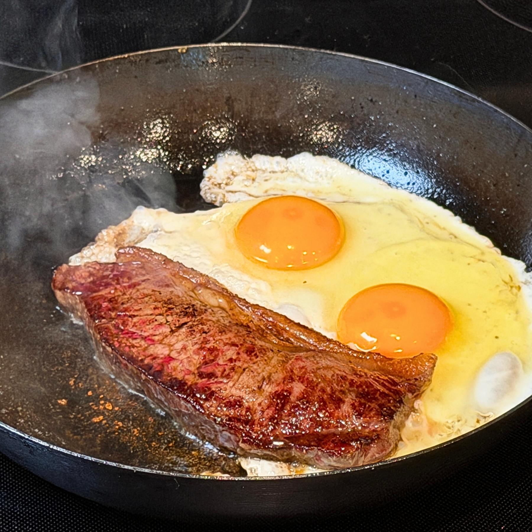 Breakfast Strip Steak searing in cast iron with two fried eggs.